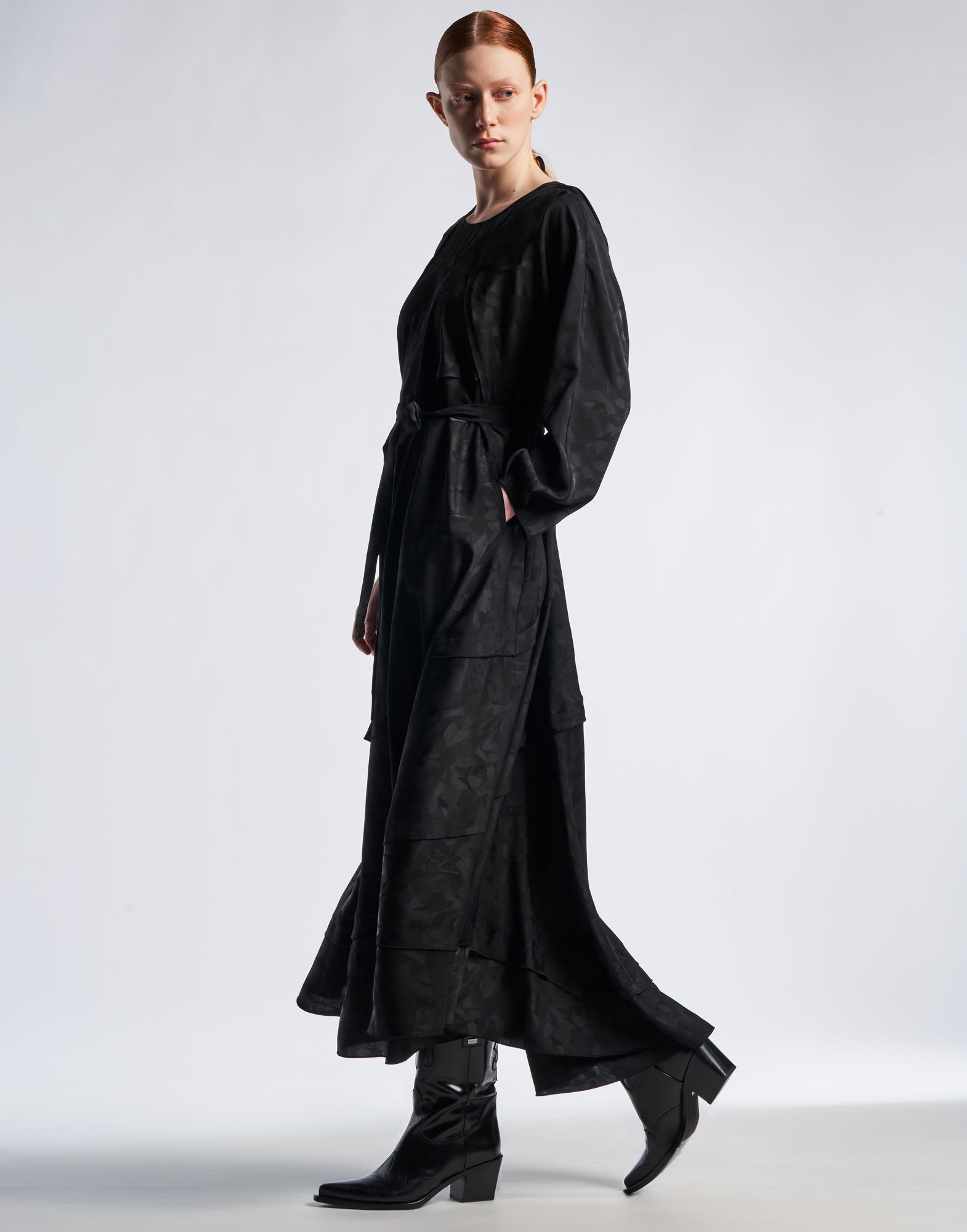 Seitenansicht: Eine Frau trägt das FAVOURITE Maxi-Kleid aus schwarzem Funktionssatin mit Damast-Blumenmuster, ausgestelltem Rock und Dolmanärmeln. Sichtbare Details wie die Paspeltasche und glatte, matte Glanz-Optik.