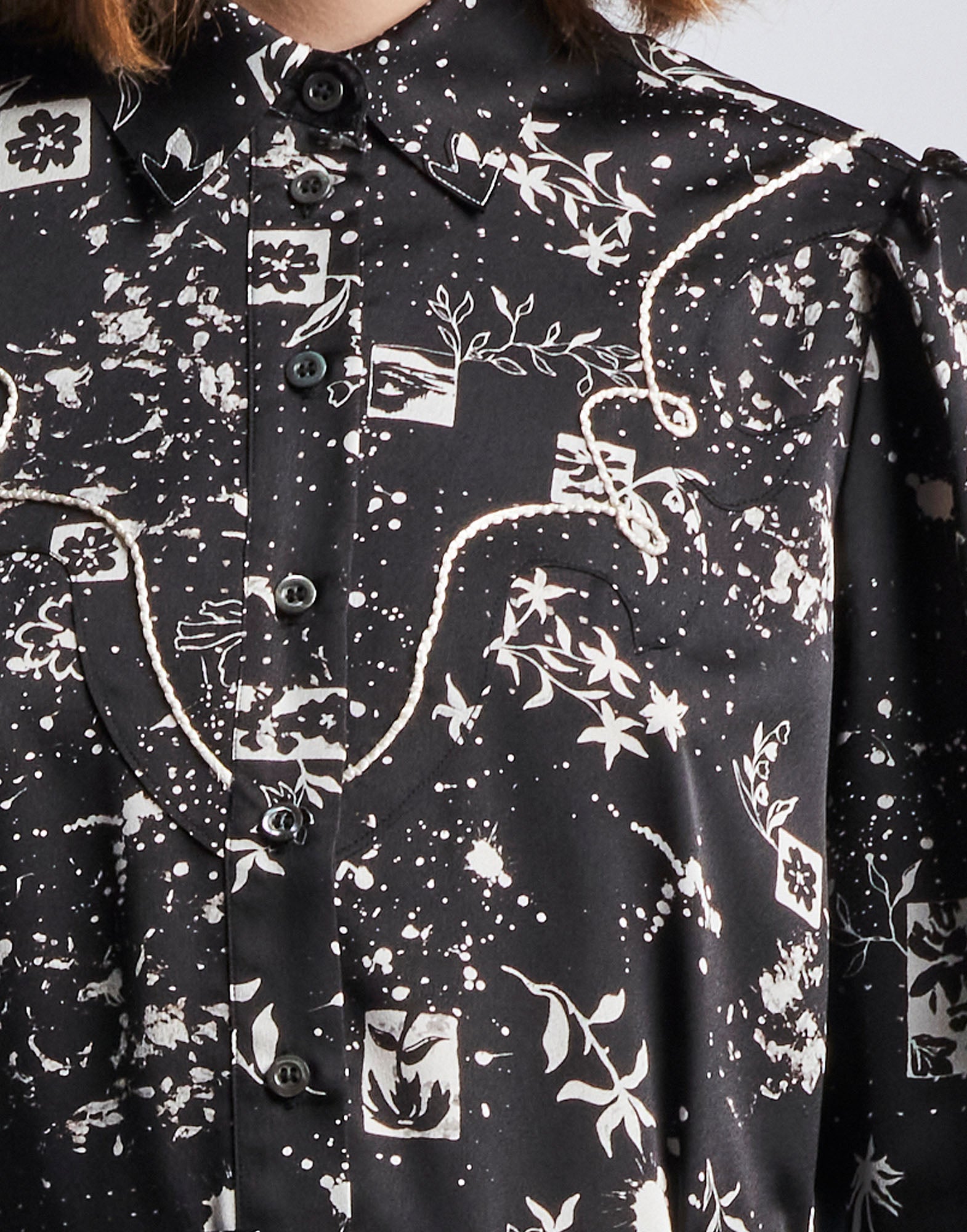 Detailansicht des Hemdkragens und der Knopfleiste des COMPELLING Hemdkleids aus schwarzem Funktionssatin mit geometrischen Blumenmustern, elfenbeinfarbenen Kordeln und Perlmuttknöpfen.