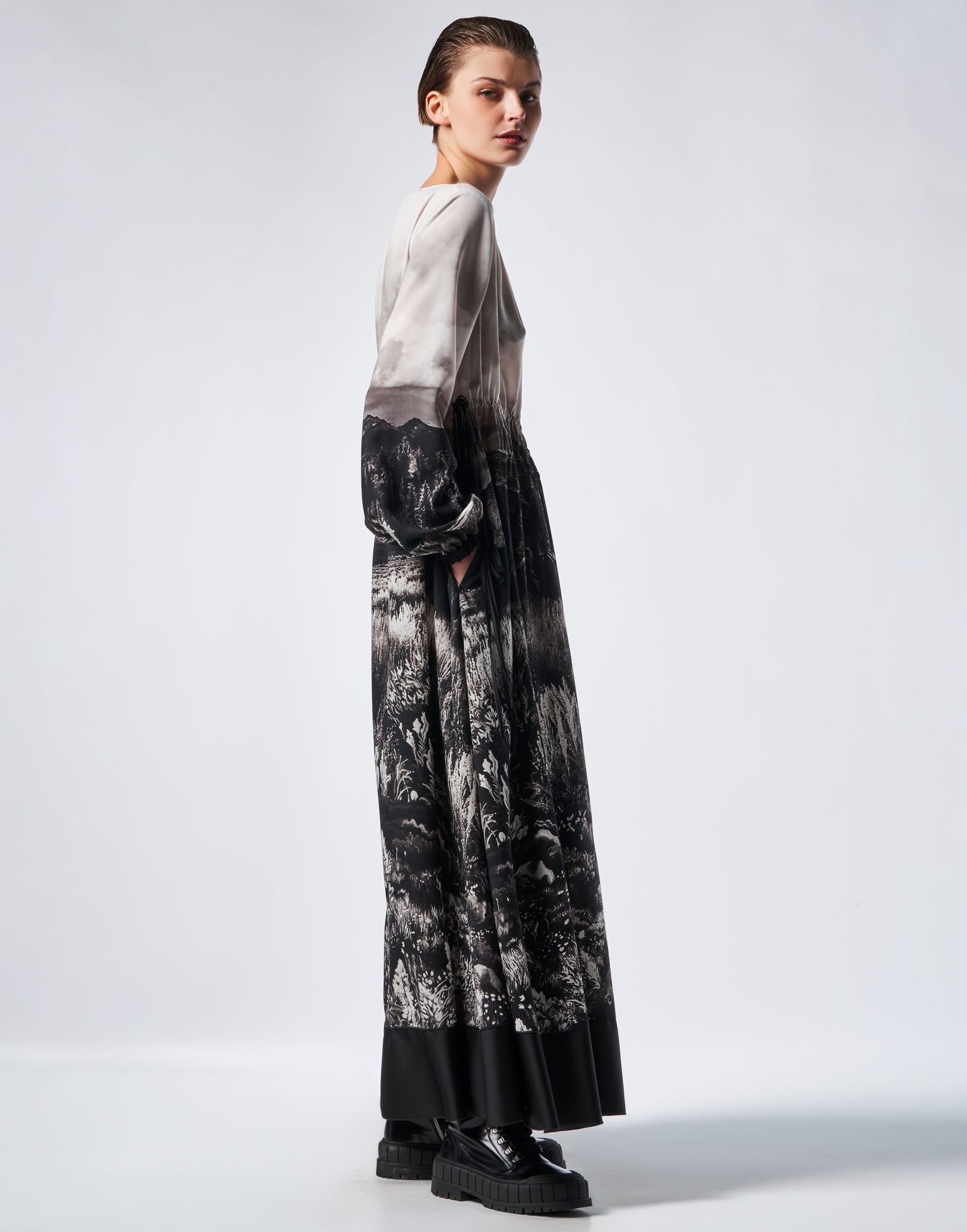 Side view of the RHYMING maxi-dress featuring black and white landscape print, long bell sleeves, and wide floor-length skirt. Model places hands in side pockets.