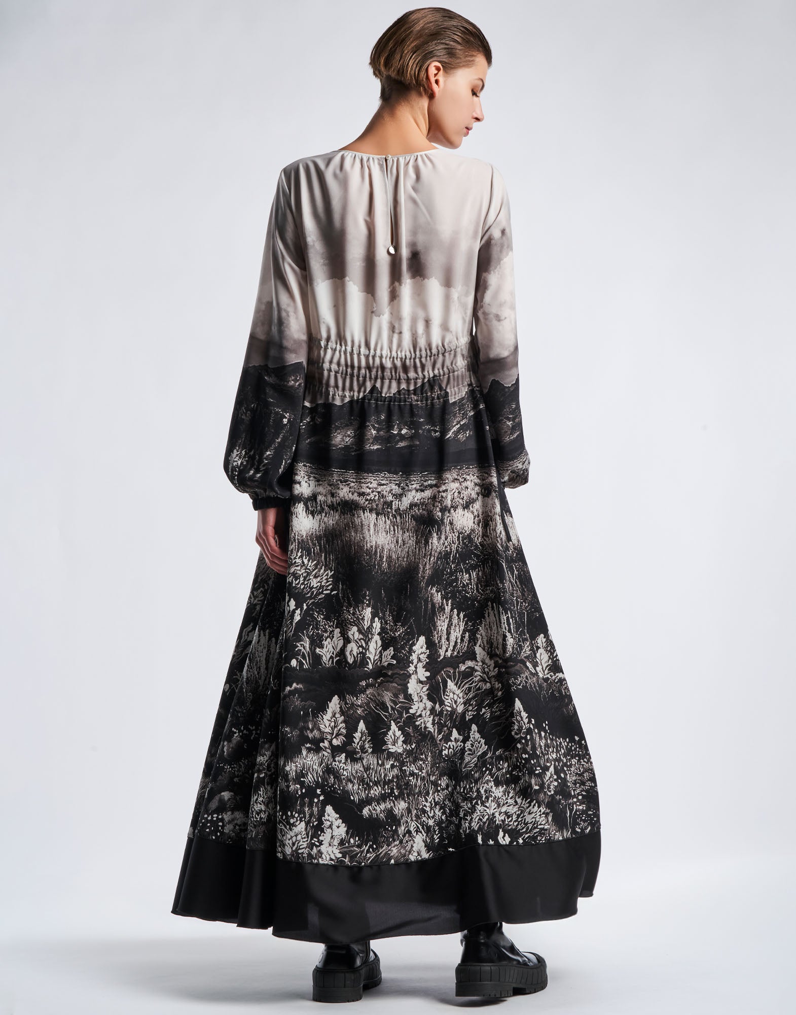 Rückansicht: Person trägt das Maxi-Kleid RHYMING mit schwarz-weißem Landschaftsdruck, weiten Glockenärmeln und bodenlangem Rock, elastischem Bund und Satinband am Saum.