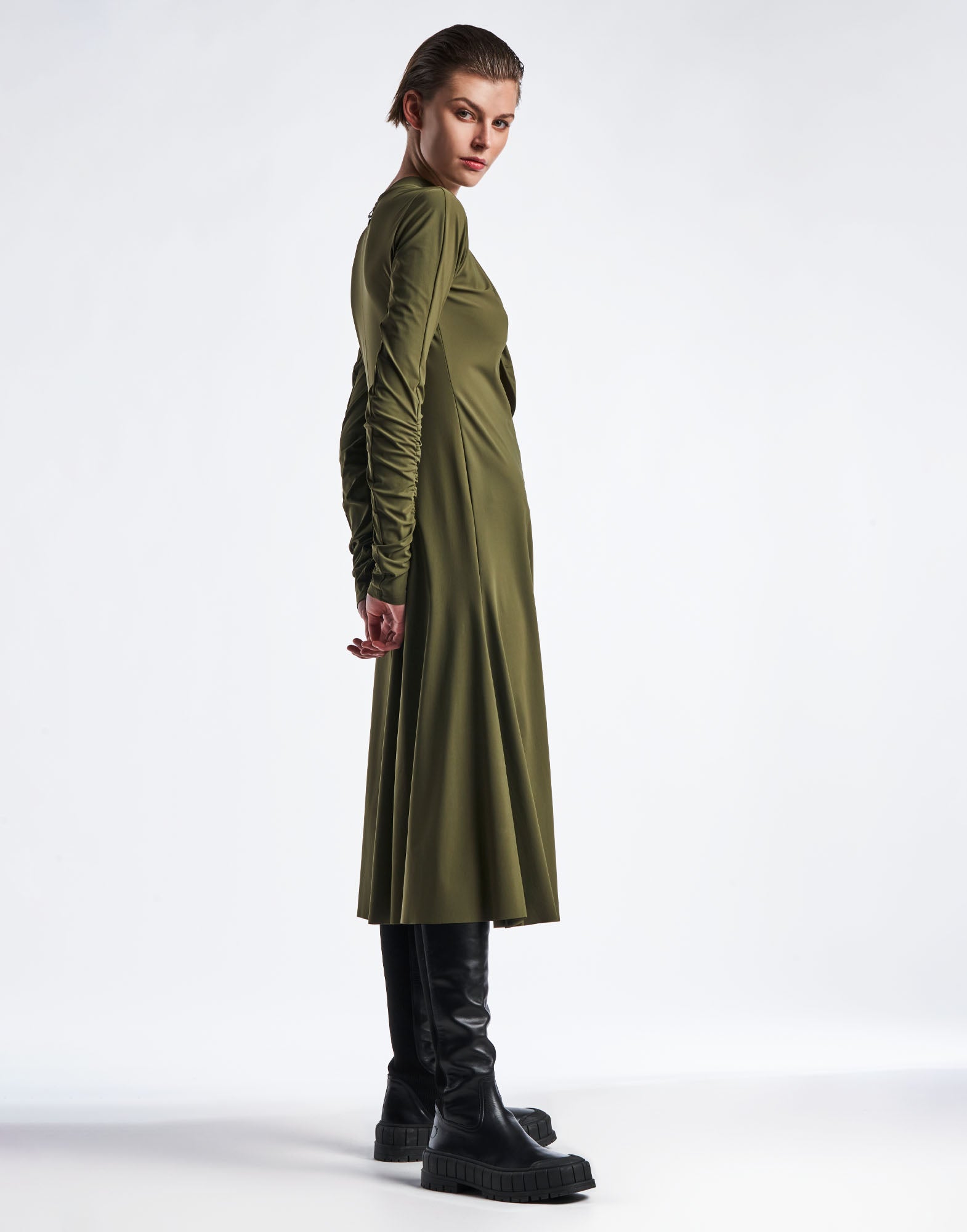 Seitenansicht eines Modells im SINCERELY Kleid, olivgrün, mit schmalen, gerafften Ärmeln, taillierter Passform und ausgestelltem Rock, kombiniert mit schwarzen Stiefeln.