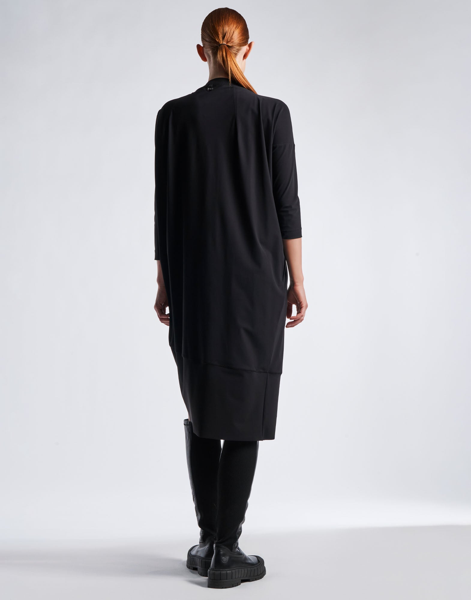 Rückansicht einer Person, die das PRELUDE Kleid trägt. Das Kleid ist schwarz, gerade geschnitten und hat dreiviertellange Ärmel. Die asymmetrische Konstruktion mit überschnittenen Schultern und konisch zulaufendem Saum erzeugt eine moderne, kokonartige Silhouette.