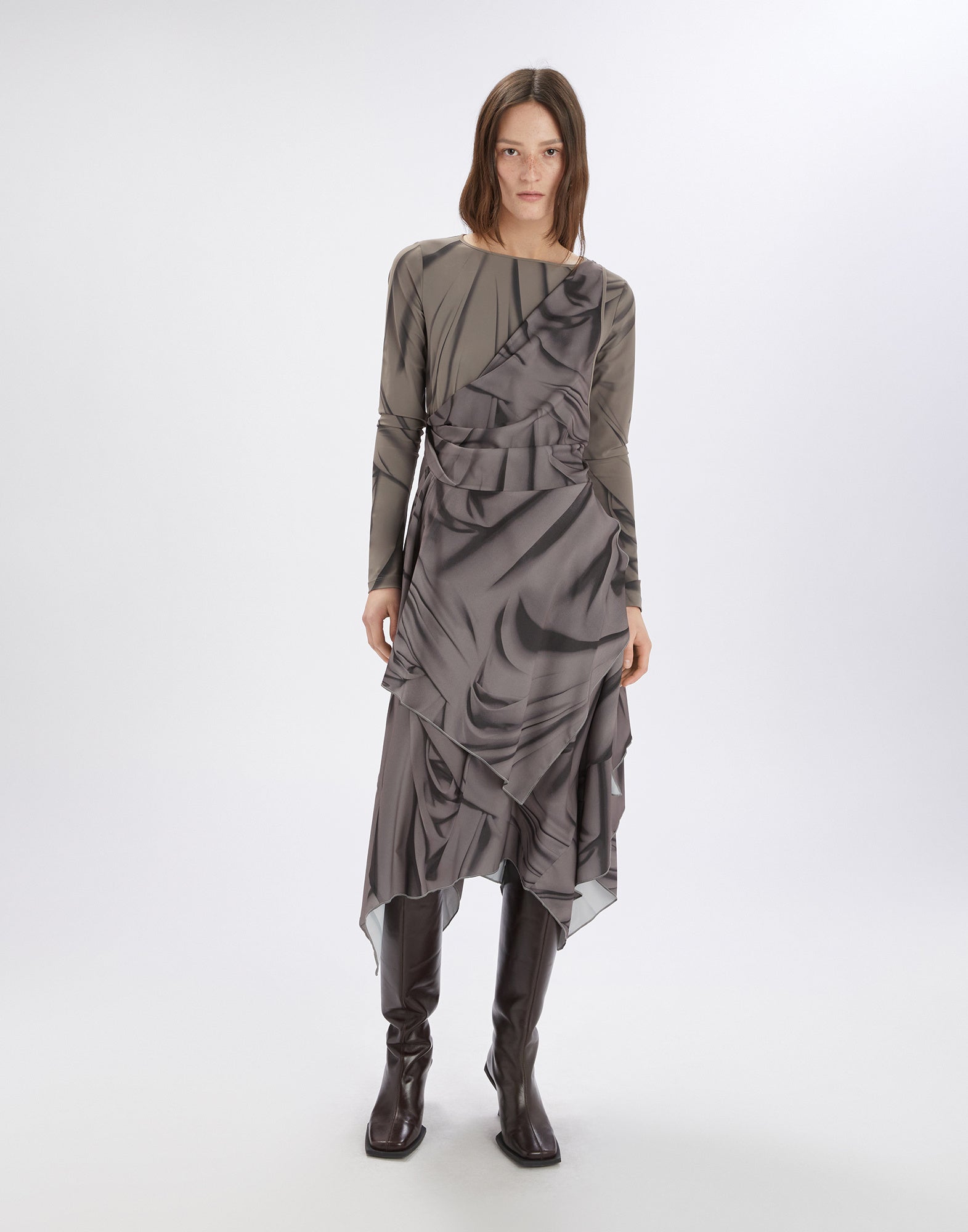 Front view: A woman wears the TROUPE dress with asymmetric hem, long sleeves, round neckline, and gray Drapery pattern. She wears knee-high boots.