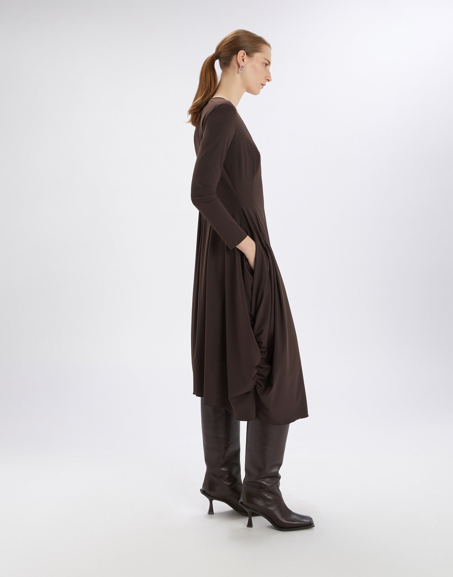 Seitenansicht eines knieumspielenden, braunen AT-LENGTH Kleids mit langen Ärmeln, V-Ausschnitt und Ballonsilhouette an einer Frau, kombiniert mit kniehohen Stiefeln.