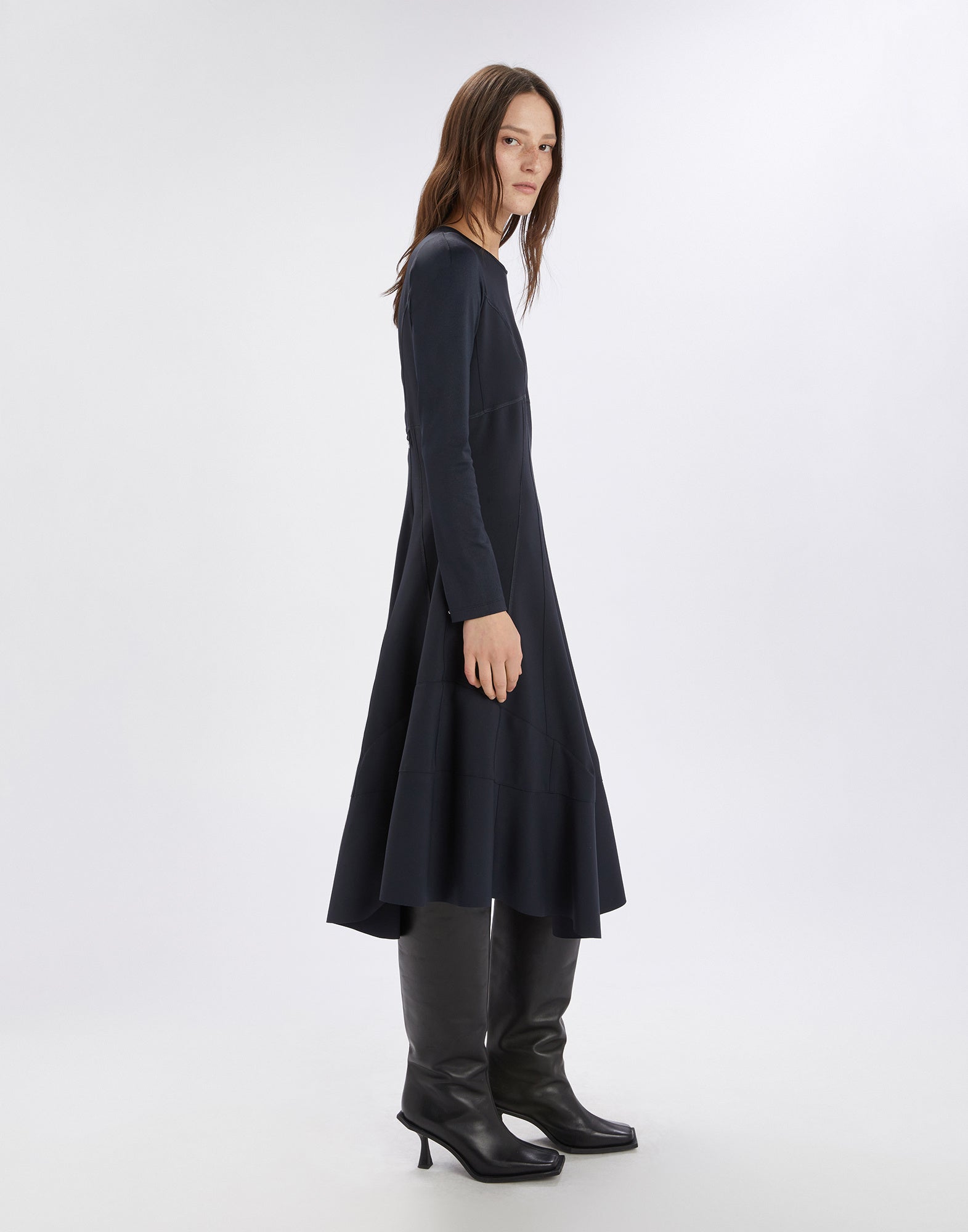 Side view of a woman wearing the SYNCHRO dress; the dress is dark, has long sleeves, visible stitched seams, and an asymmetric hem.