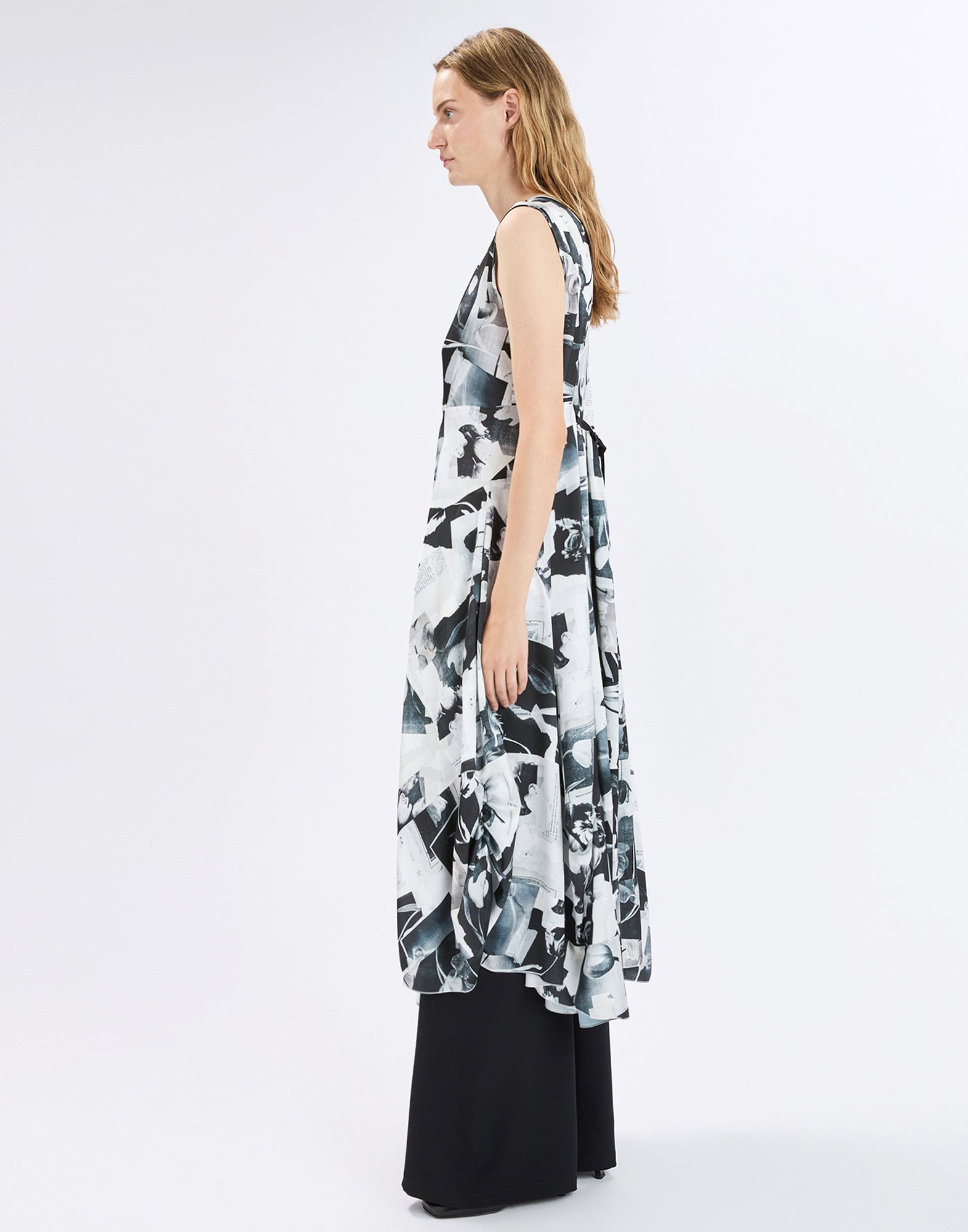 Side view: model wears the sleeveless black-and-white printed SPARKY dress with an asymmetric, voluminous hem over black trousers against a light studio background.