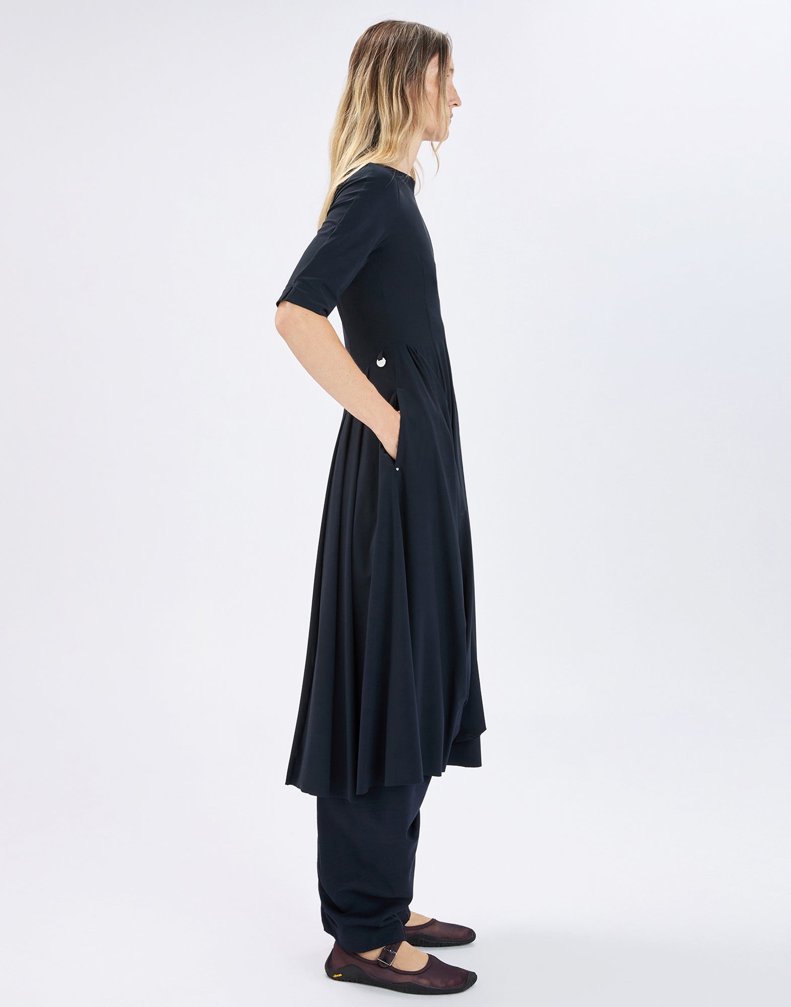 Side view: model wearing the ankle-length, dark blue NEW MESSENGER dress with elbow-length sleeves, slightly flared irregular hem and side pocket, paired with slim pants.