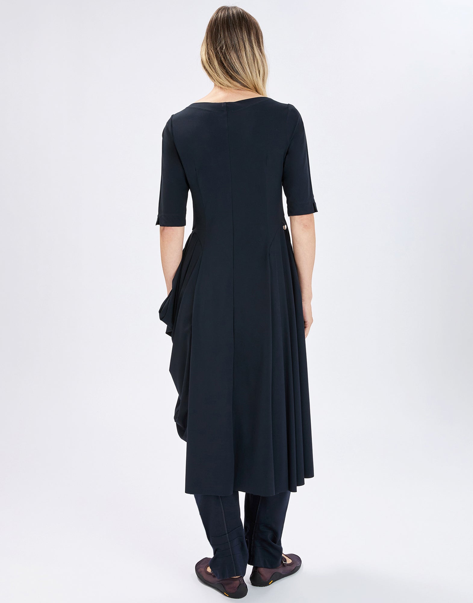 Rear view: Person wearing dark blue NEW MESSENGER dress with elbow-length sleeves, asymmetrically draped skirt and longer back, paired with dark pants and flat shoes.