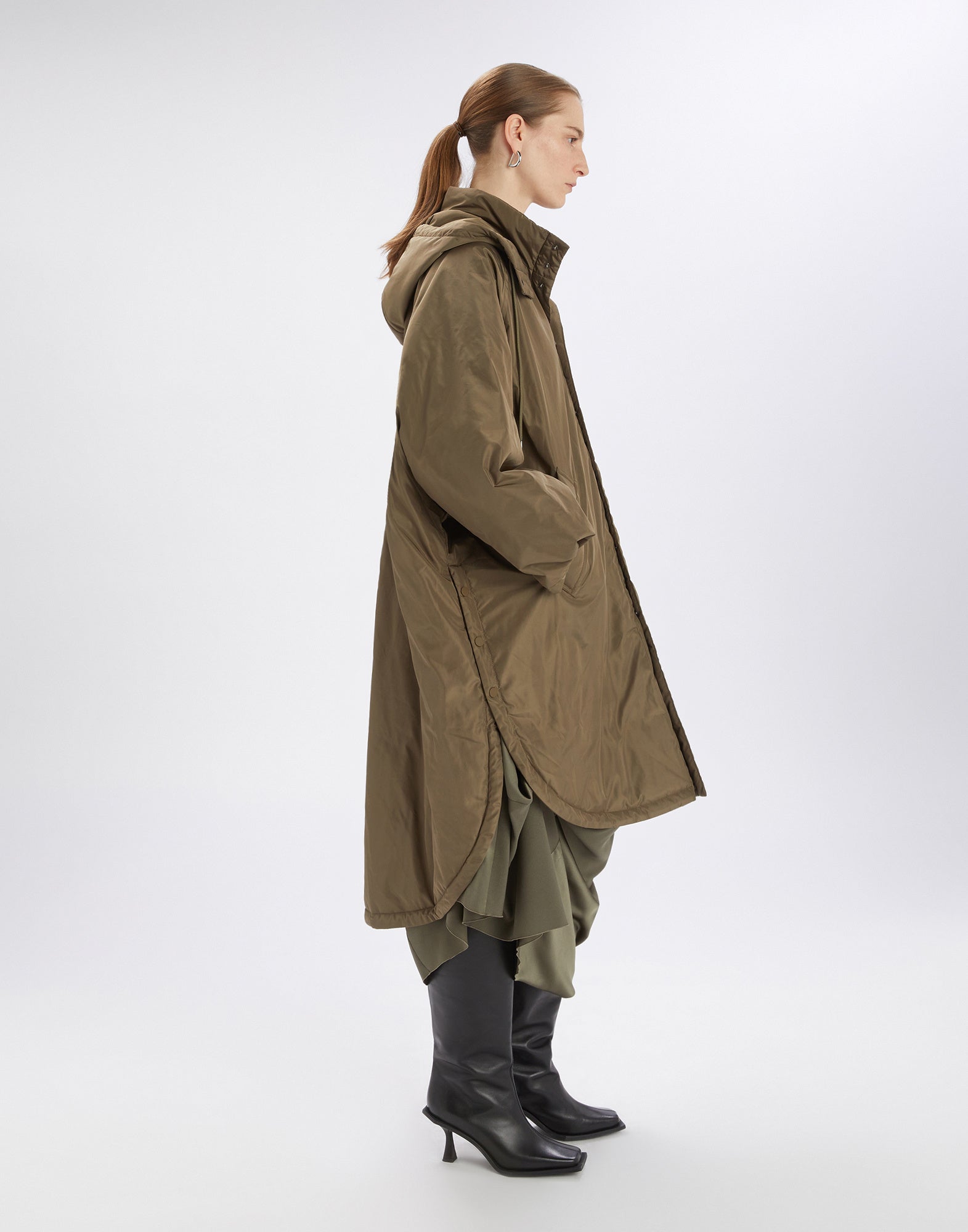 Seitenansicht eines Models, das den Parka COFFER trägt. Der Mantel ist oversized, wasserabweisend, olivfarben, mit Kapuze, seitlichen Knöpfen und weitem Kimono-Ärmel.