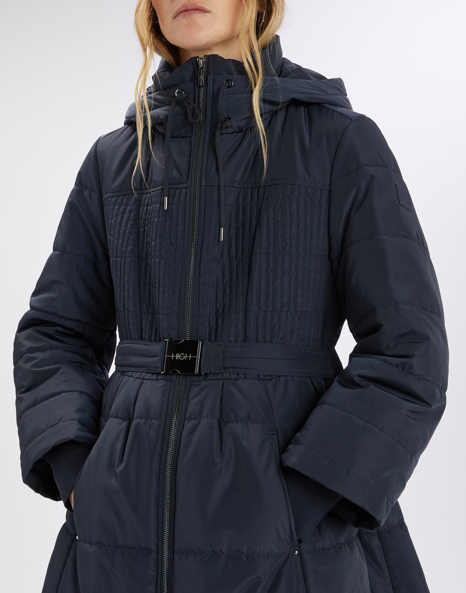 Front view of a person wearing the SCUTTLE down jacket with embroidered quilting, elastic waist belt, hood with drawstring, and visible zipper.
