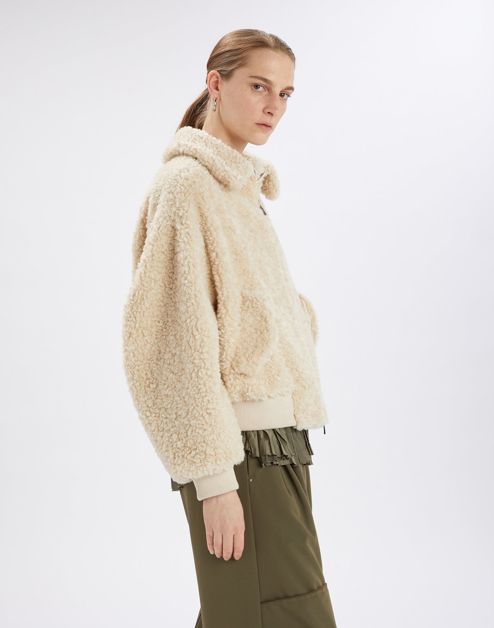 Seitenansicht einer Person, die die RULER Sherpa-Bomberjacke aus cremefarbenem Boucle-Fellstoff trägt. Jacke mit umgeschlagenem Kragen, Reißverschluss und Oversize-Passform.