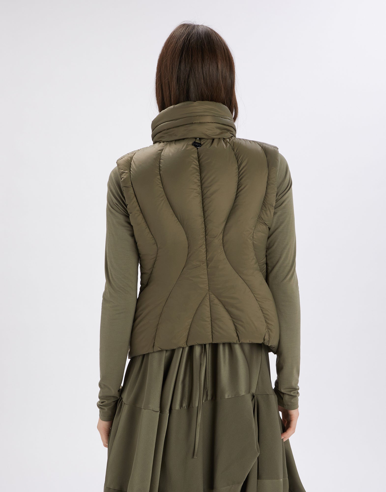 Rückansicht der Weste SPIFFY in Olivgrün, getragen von einer Frau. Die Weste zeigt geschwungene Nähte und einen hohen Stehkragen mit integrierter Kapuze.
