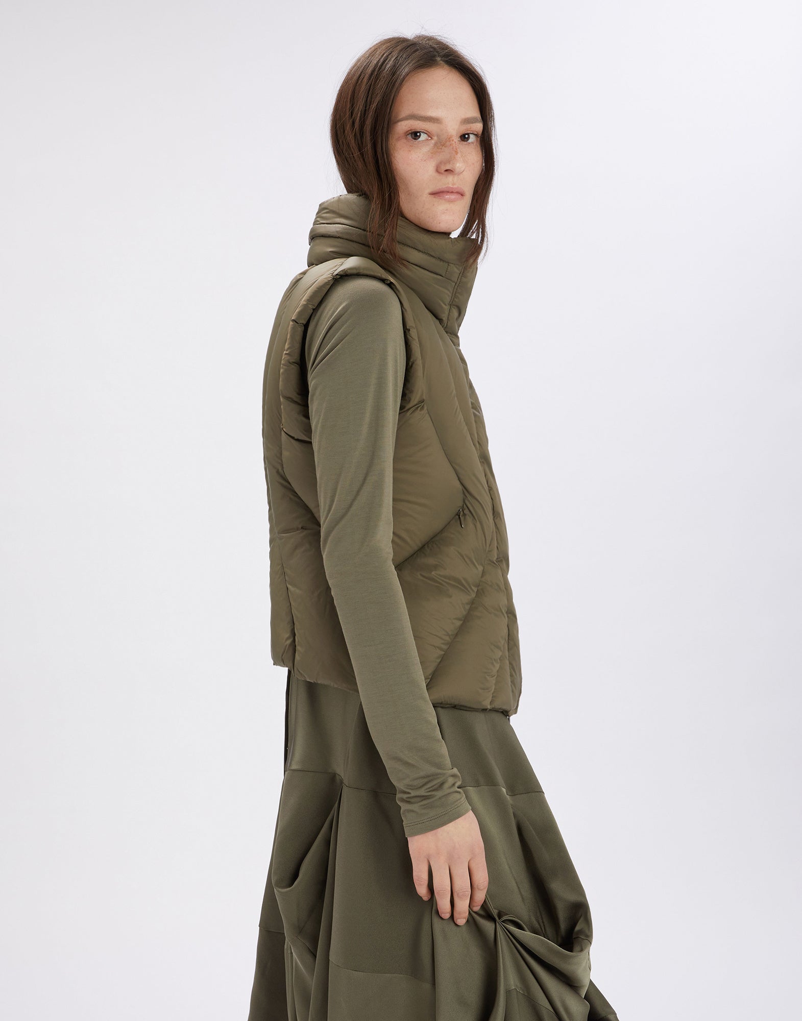 Seitenansicht einer Frau, die die ultraleichte, olivgrüne SPIFFY Weste mit hohem Stehkragen und integrierter Kapuze trägt. Die geschwungenen Nähte und Diagonalreißverschlusstaschen der Daunenfüllung sind sichtbar.