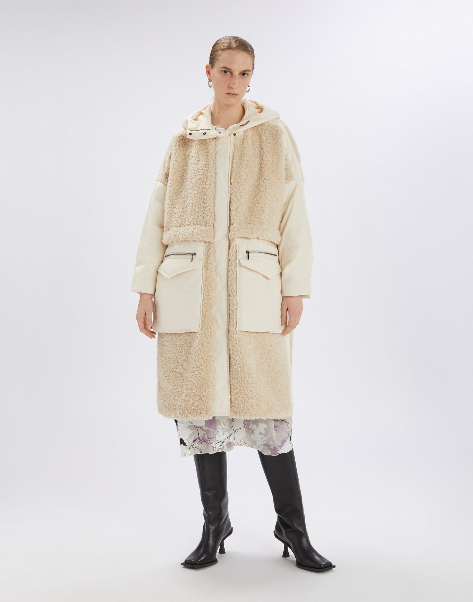 Front view of a person wearing the cream-colored ANNOTATE coat made of sherpa fabric and nylon, featuring a hood, large front pockets, and knee length.