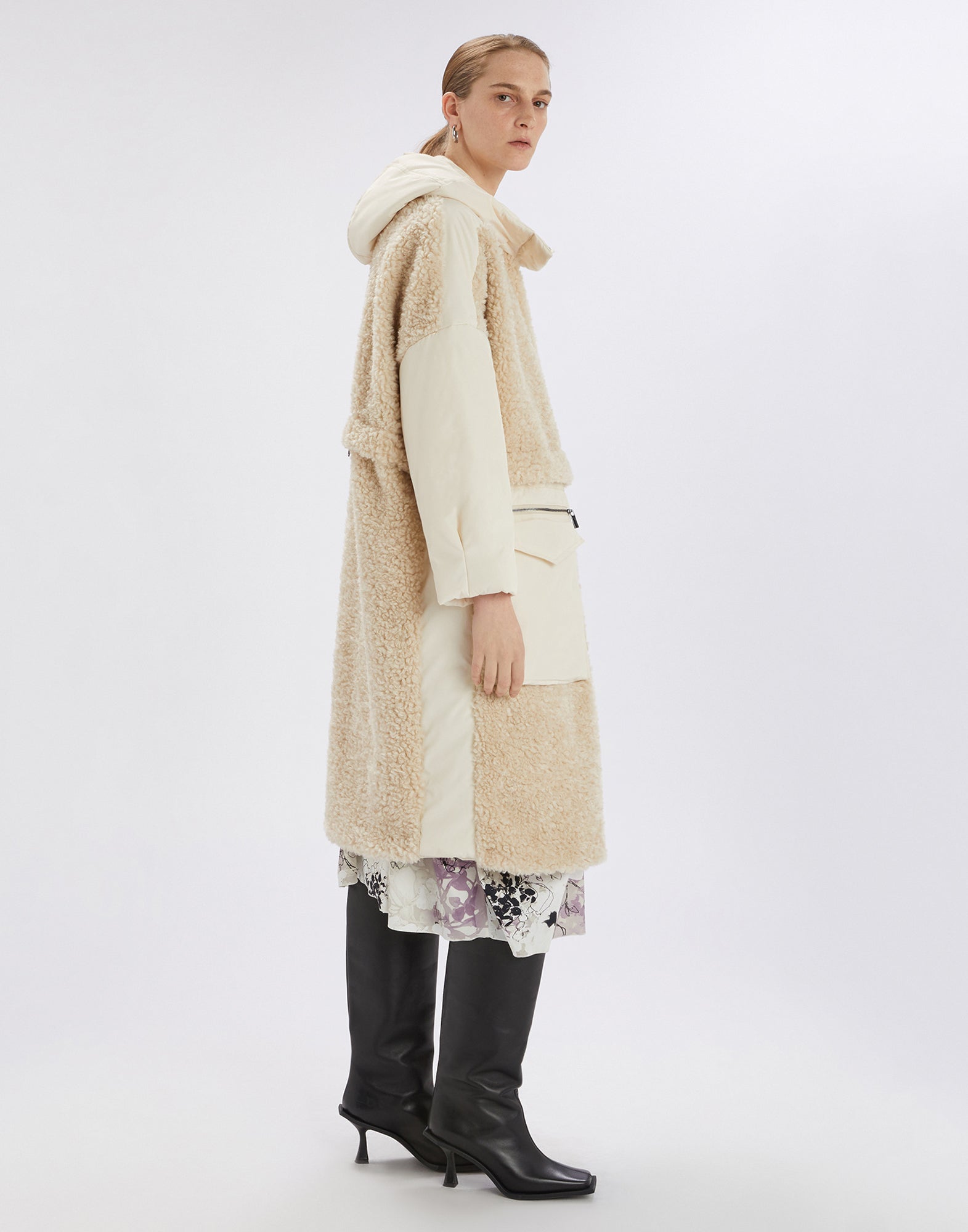 Side view of the ANNOTATE coat, knee-length with beige sherpa look, nylon inserts, hood, and large front pockets. Worn by a person with boots.