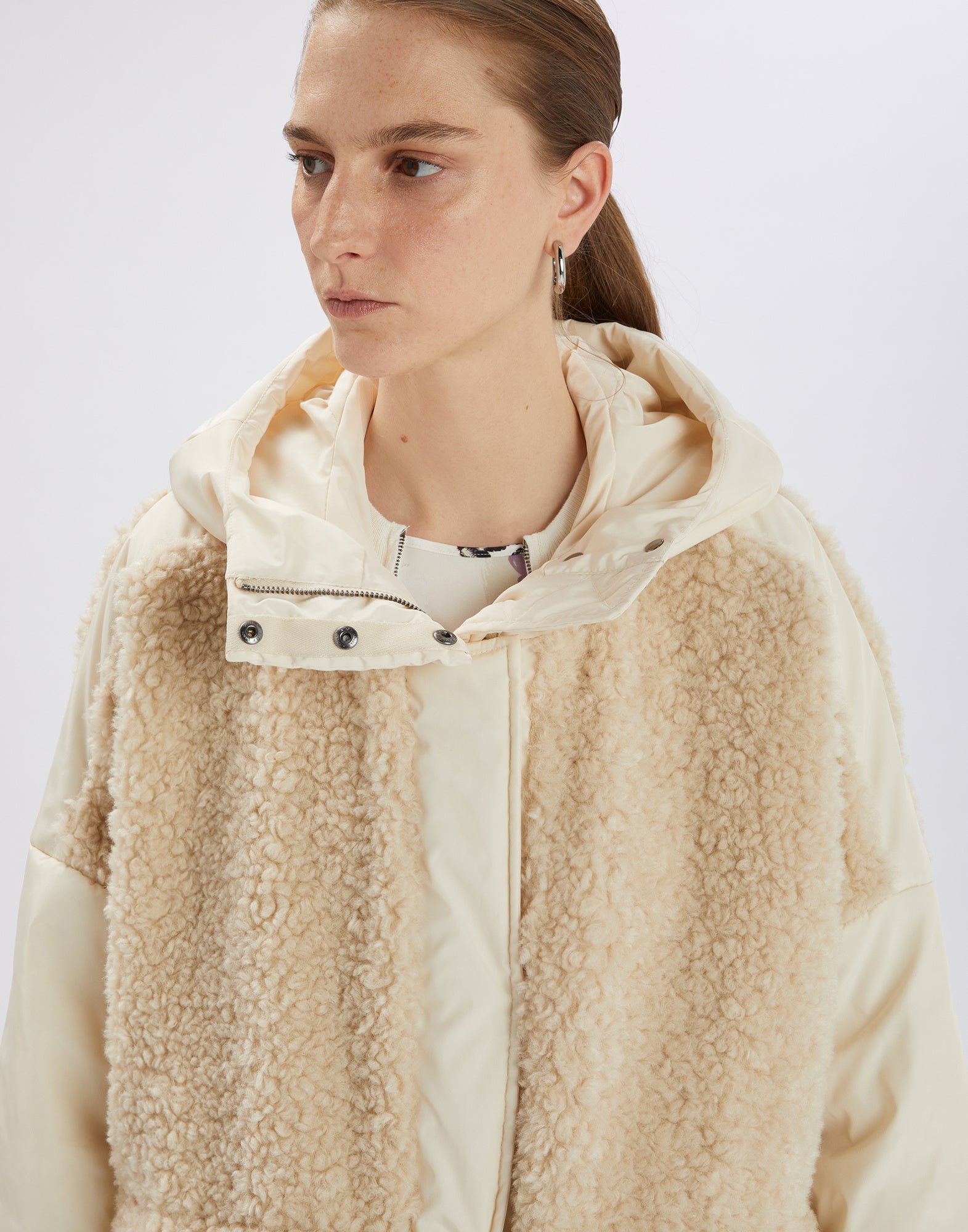 Front view of the ANNOTATE coat, worn by a woman. The coat is made of cream-colored sherpa fabric with nylon details, hood, and large front pockets.