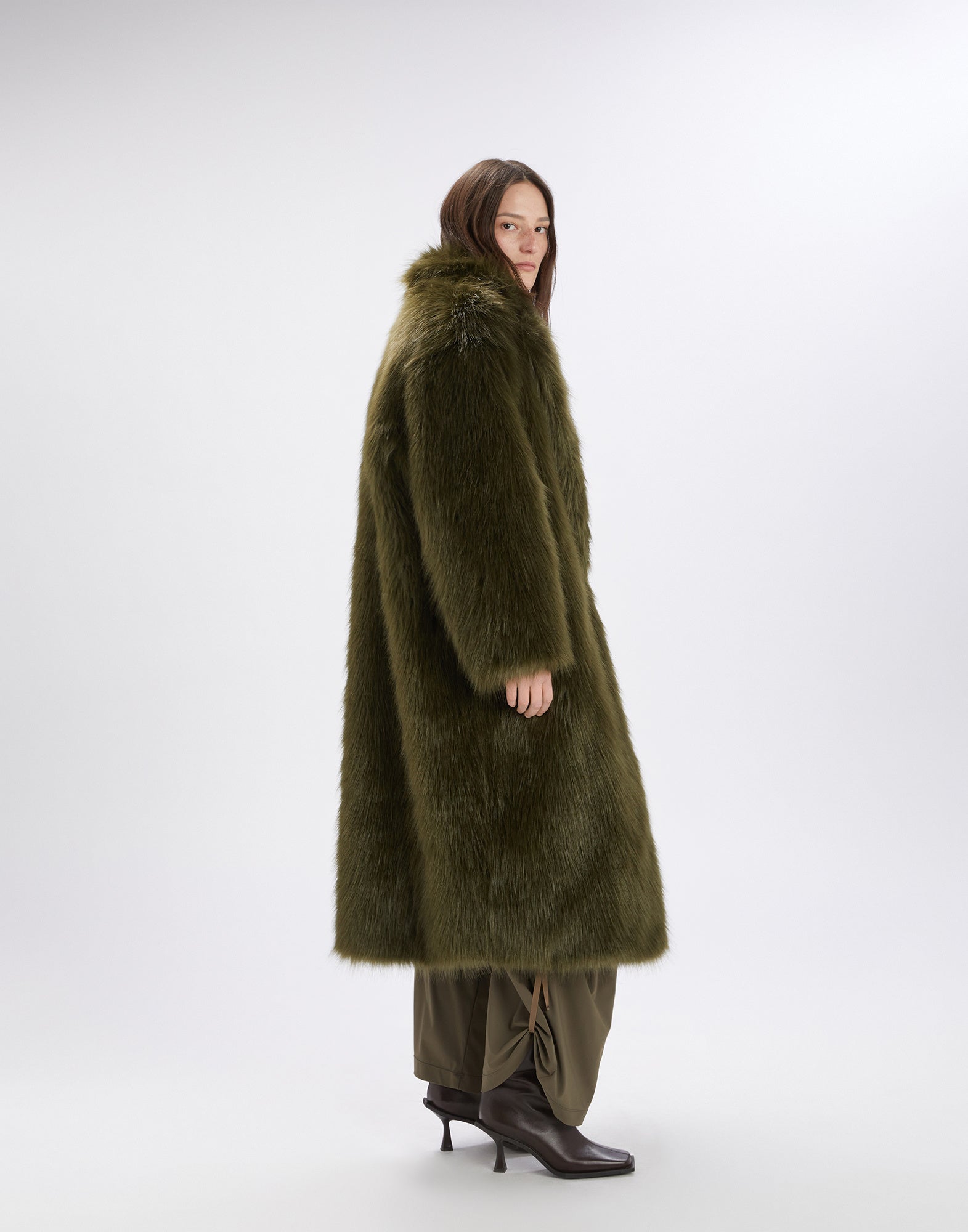 Seitenansicht: Eine Frau trägt den Kunstpelzmantel CROSSING in olivgrün mit Maxi-Volumen, Reverskragen und Länge bis knapp über die Knöchel in schlichter Studioumgebung.