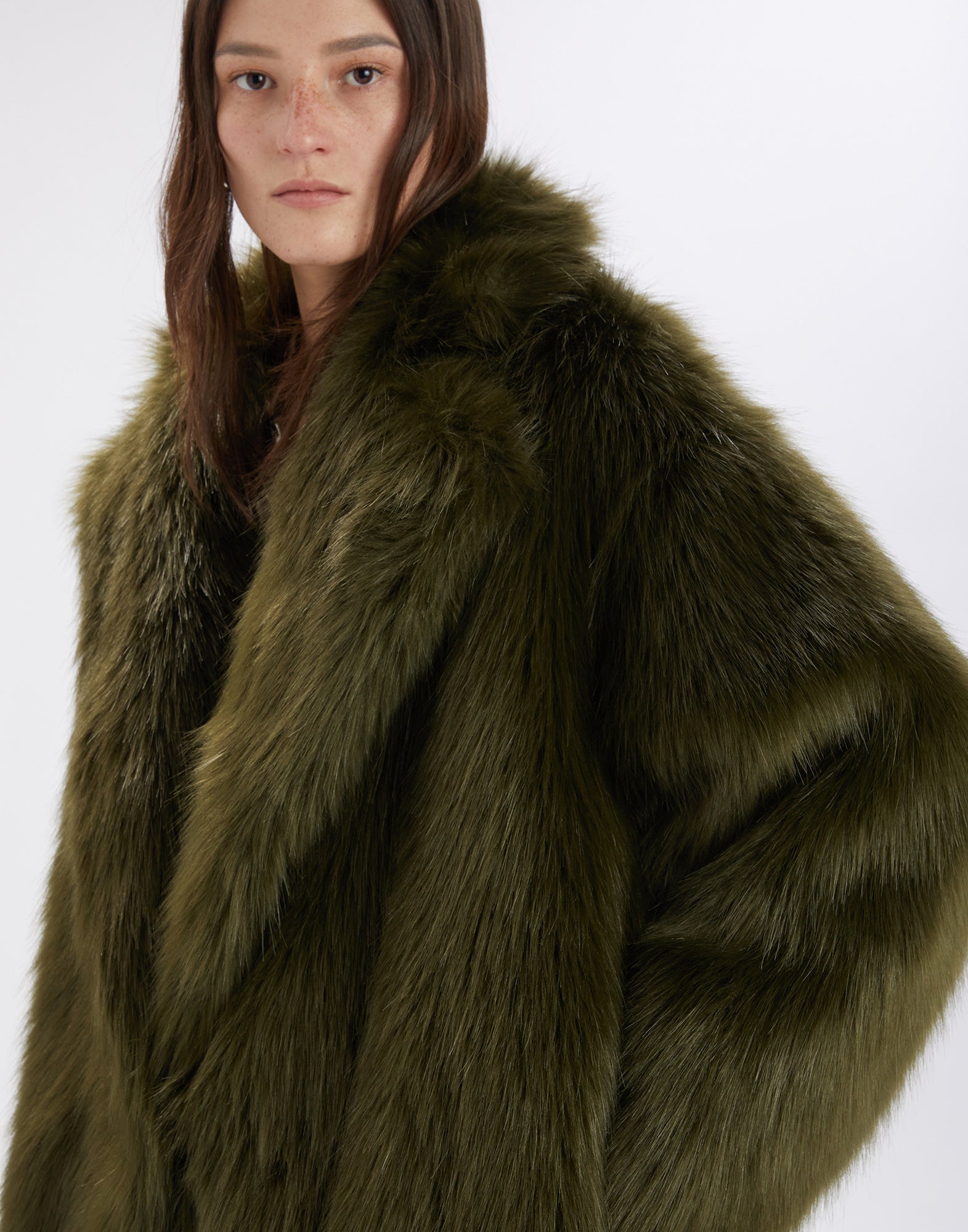 Frontalansicht einer Frau, die den CROSSING Kunstpelzmantel in olivgrüner Farbe trägt; der Mantel hat ein voluminöses Maxi-Design und breite Revers.