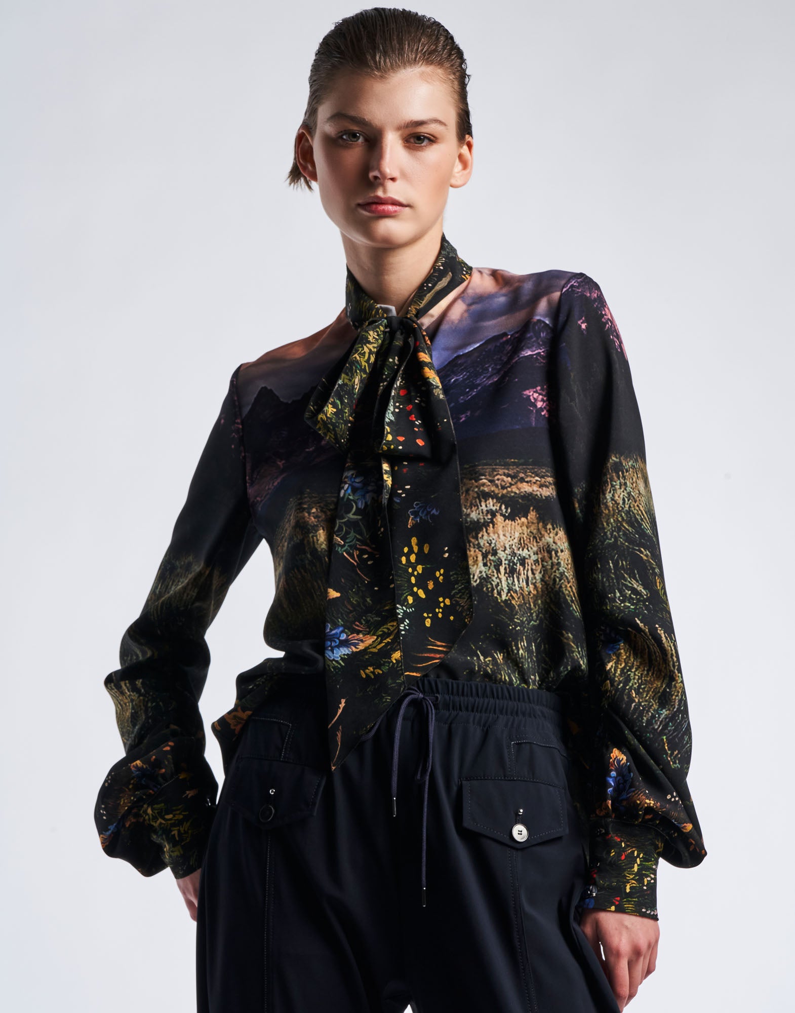 Frontalansicht: Person trägt AMAZEMENT Bluse aus Funktionskrepp mit mehrfarbigem Wyoming-Druck, Stehkragen mit Schleife, langen Ärmeln und Dreiknopfmanschetten.