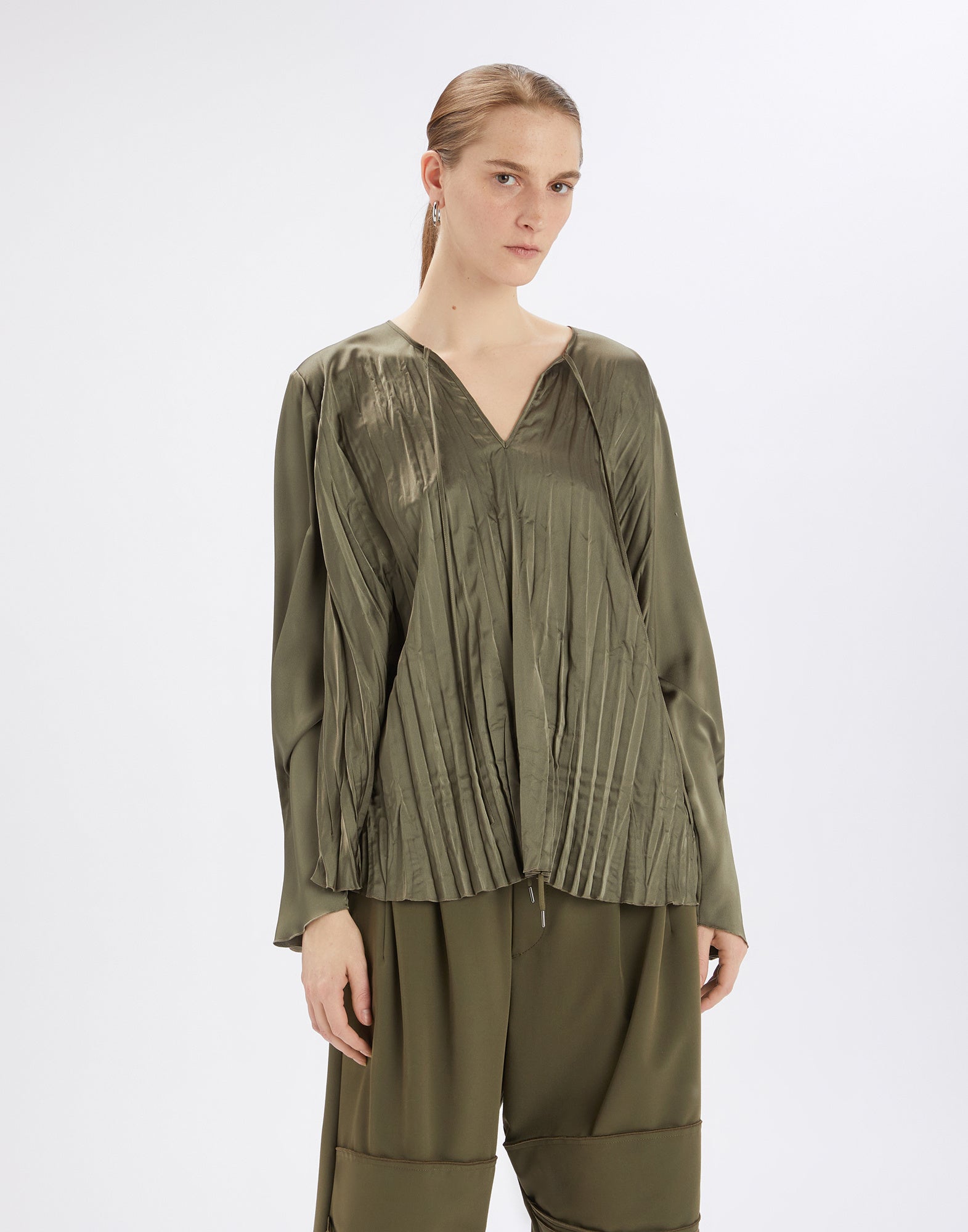 Frontalansicht einer Person, die die ONTOP Bluse trägt; olivgrüner, glänzender Satin mit Plisseeeffekt, weitem Rundhalsausschnitt und langen, leicht gerafften Ärmeln.