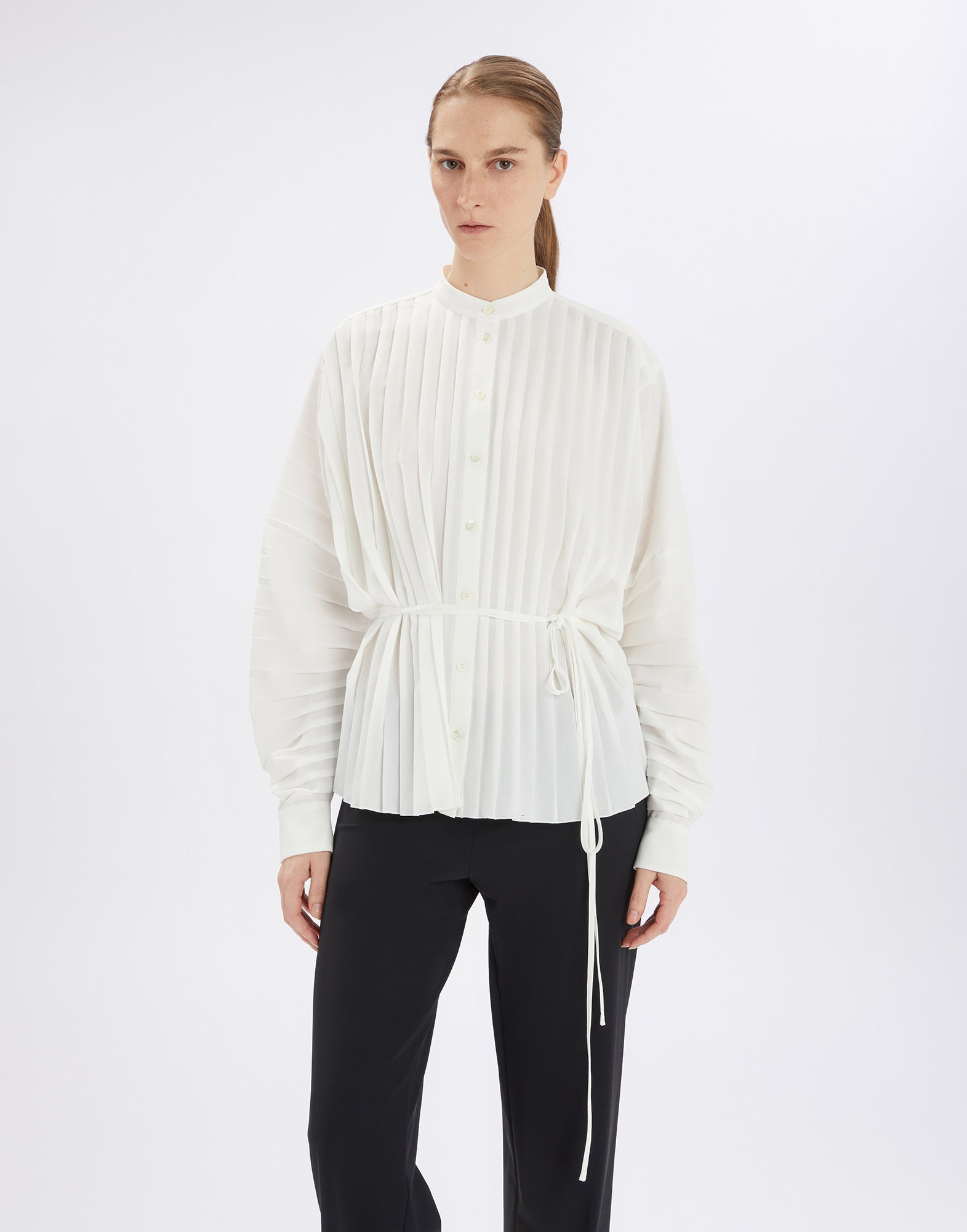 Frontalansicht der Bluse SLIM, getragen von einer Person. Die Bluse ist weiß, plissiert, hat einen Mao-Kragen, Knopfleiste, lange Ärmel und einen Kordelzug in der Taille.