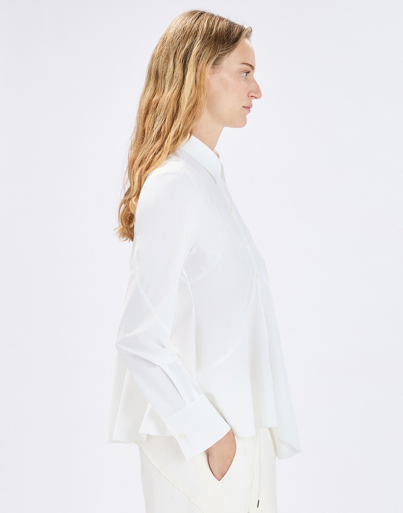 Side view: model wearing the white DEBONHAIR blouse with notched collar, long button placket, long sleeves with wide cuffs, and a slightly flared, irregular hem, styled with light-colored pants.