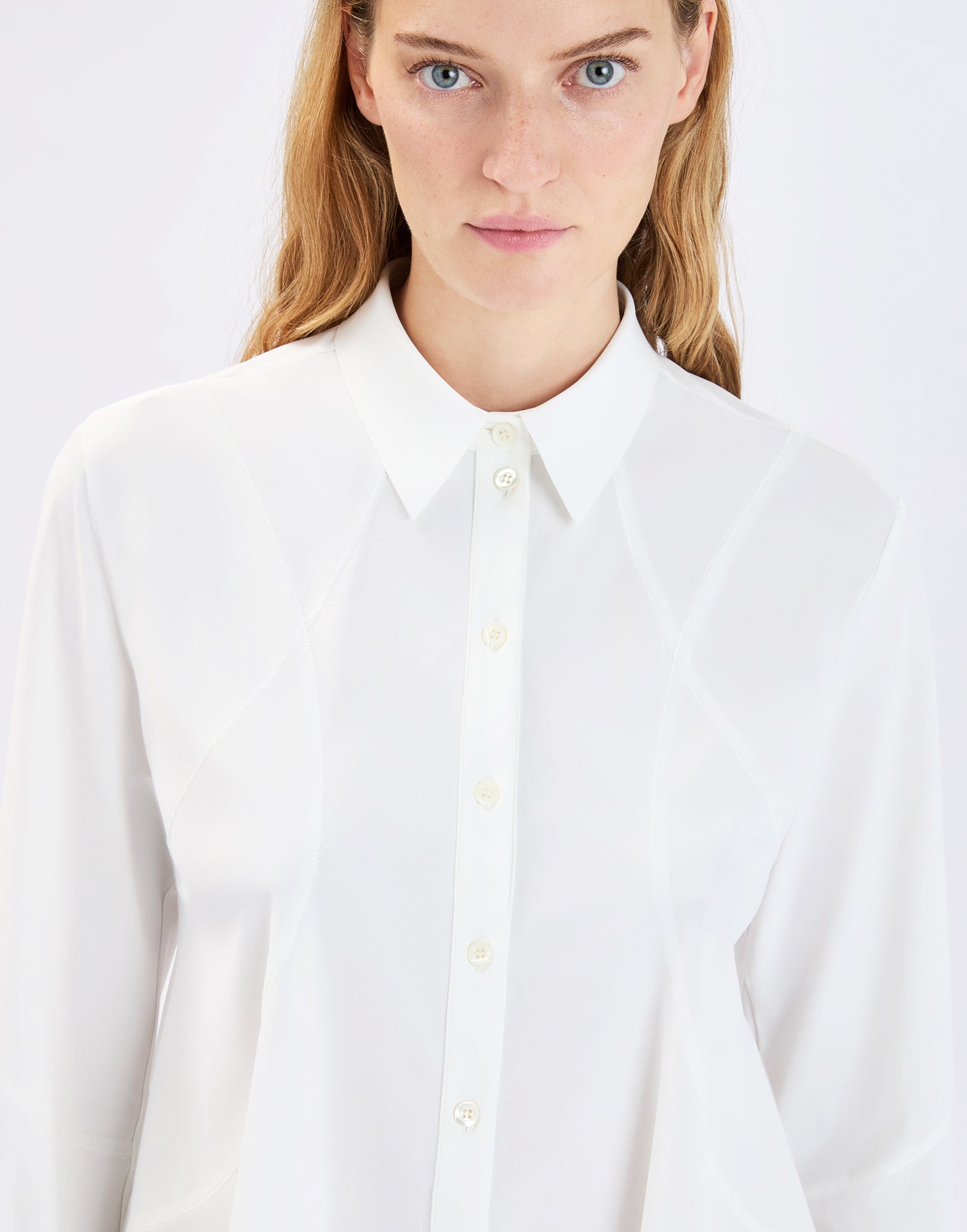 Front view: Person wearing the white DEBONHAIR blouse with notched collar, full button placket, and subtly visible exposed seams that create a slim, slightly flared silhouette.