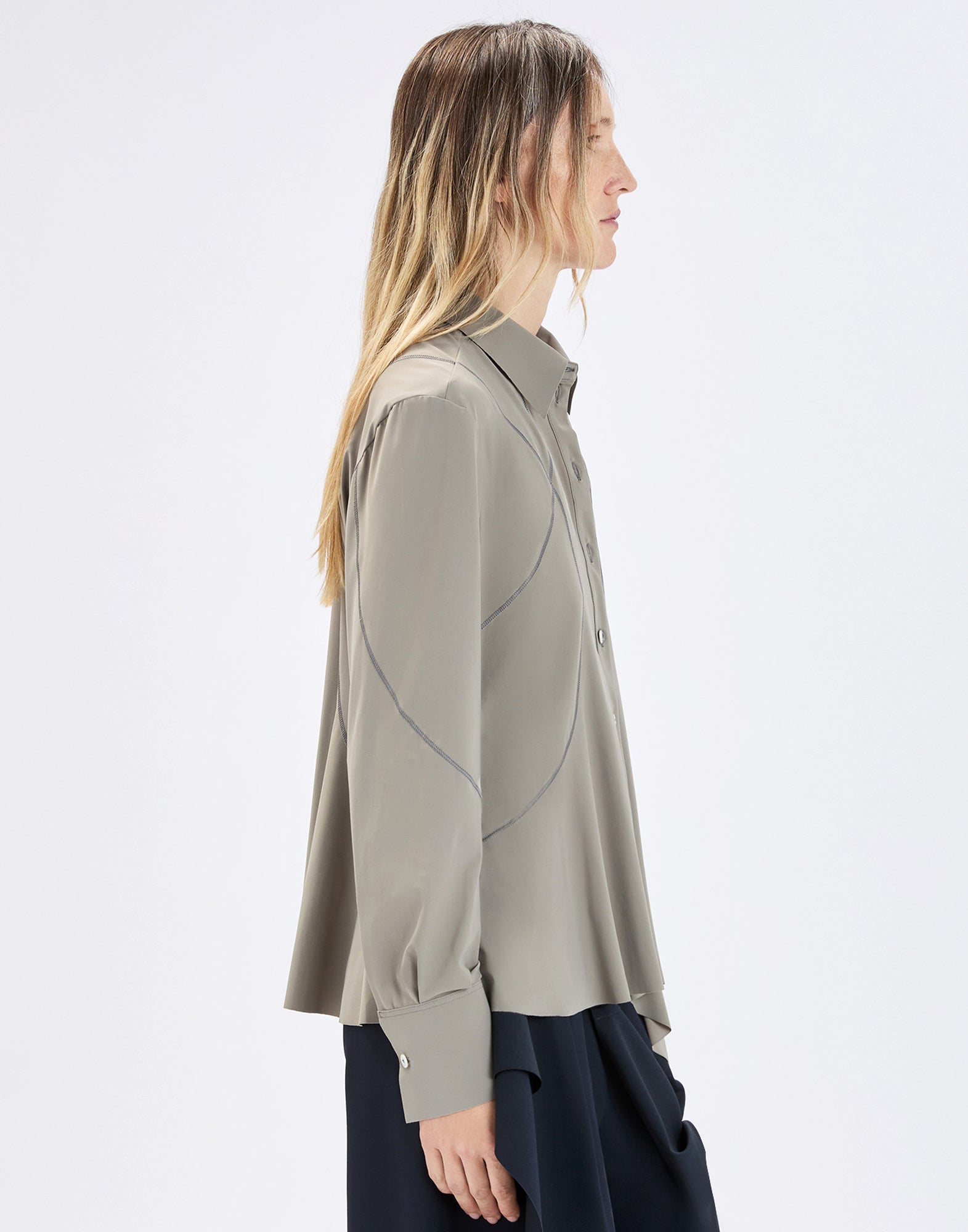 Side view: model wearing the loose-fitting DEBONHAIR blouse in light khaki with visible curved seams, classic collar, button placket, and long sleeves with wide cuffs.