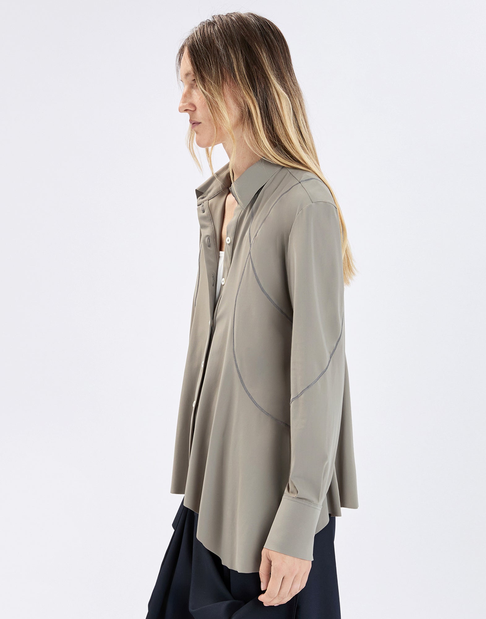 Side view: person wearing loose-fitting DEBONHAIR blouse in olive gray with notched collar, button placket, visible curved seams, and long sleeves with wide cuffs over a dark skirt.