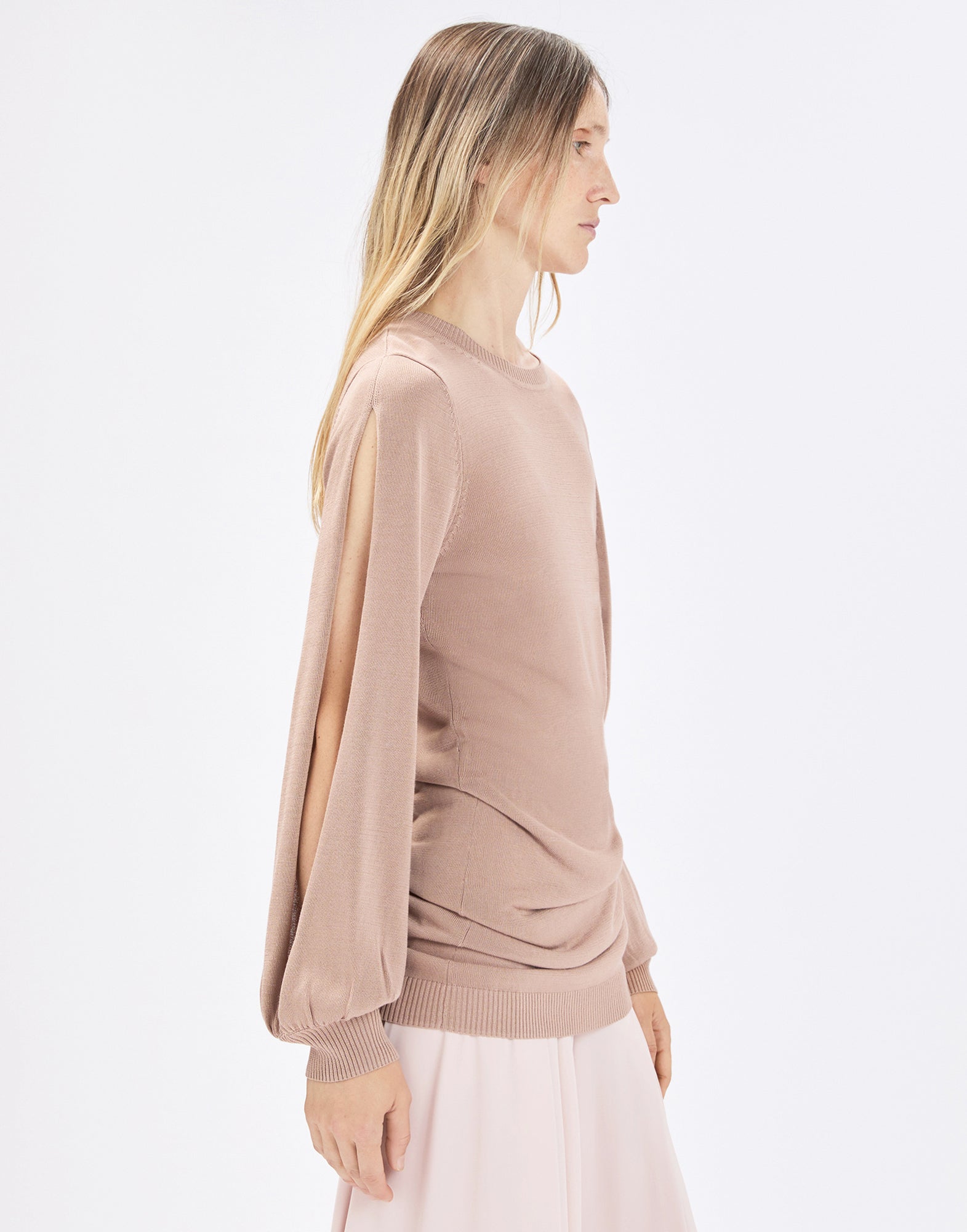 Side view: person wearing the beige SANDUST knit crew-neck top with side-slit balloon sleeves and light pleating at the chest, paired with a light-colored skirt.
