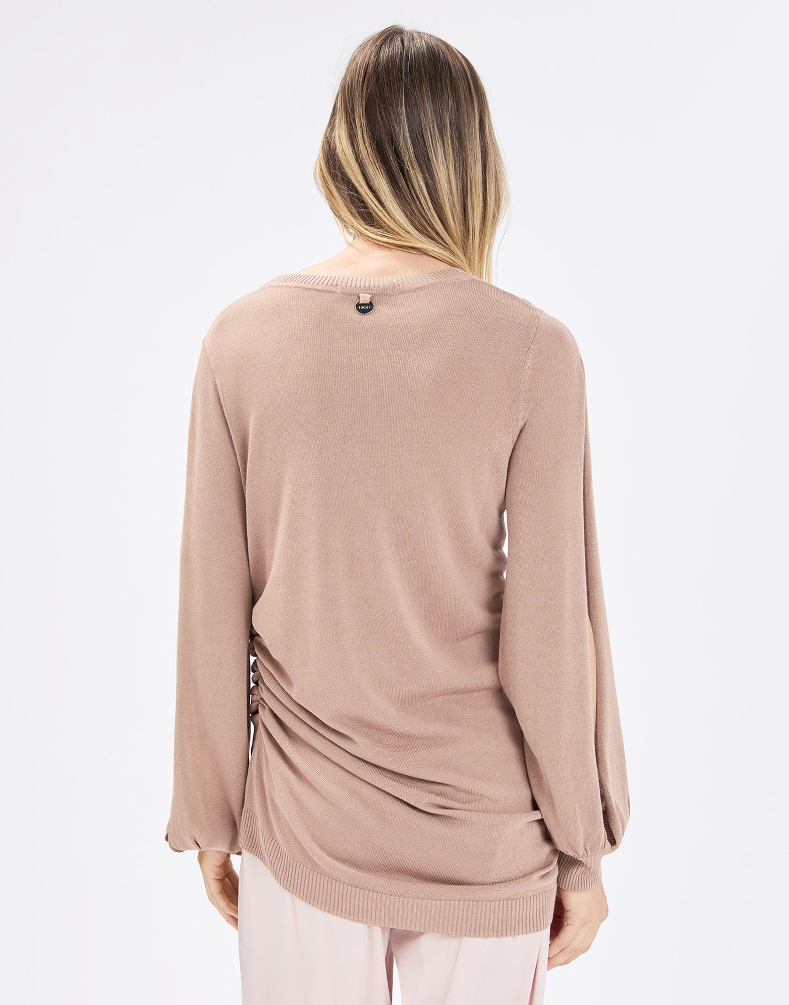 Back view: model wearing the beige SANDUST knit top with long sleeves, crew neckline, and ruched hip area at the side, loosely draping over light-colored pants.