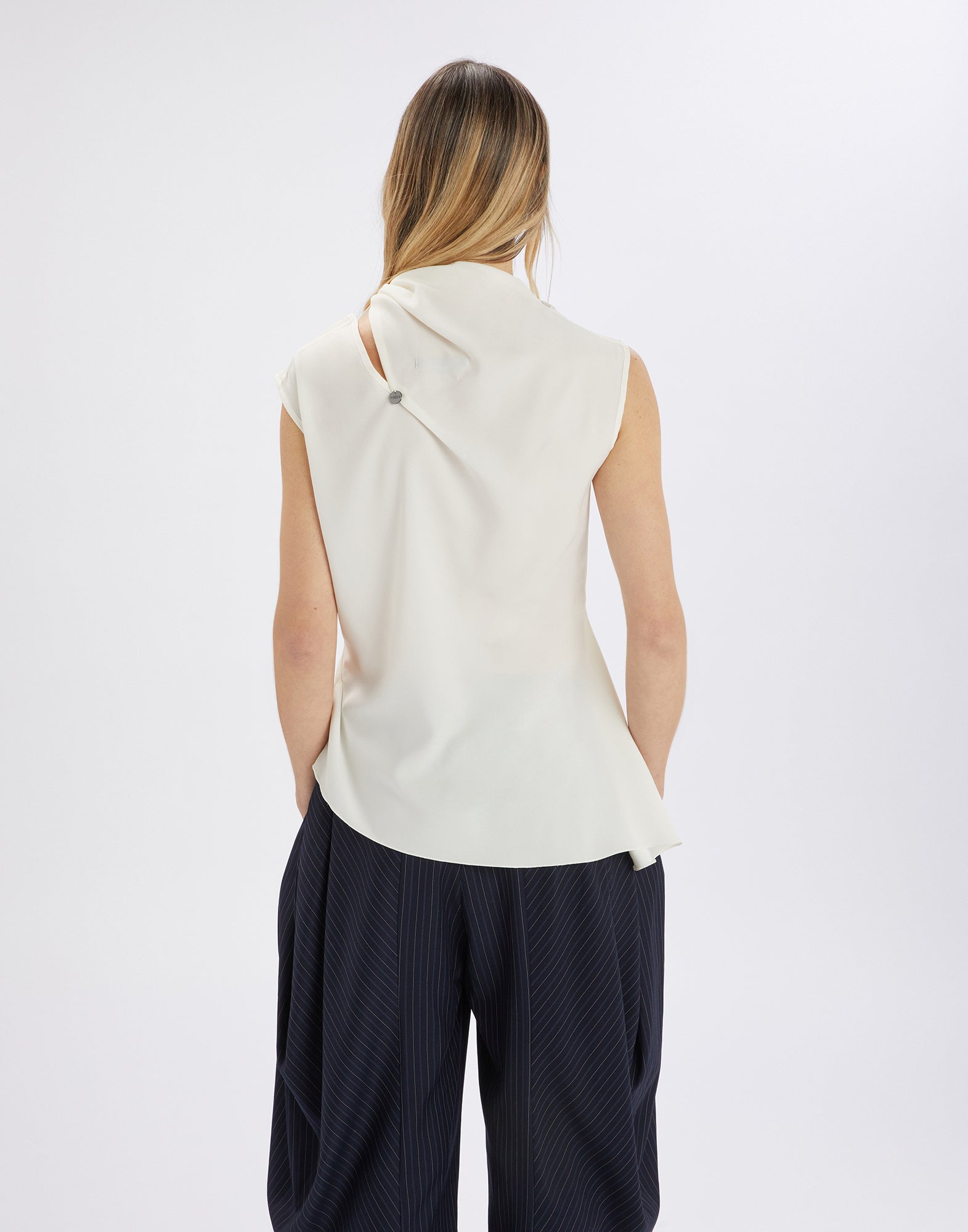 Rear view of a woman wearing the sleeveless WALTON top in flowing cream satin; asymmetric hem and side jabot collar visible.
