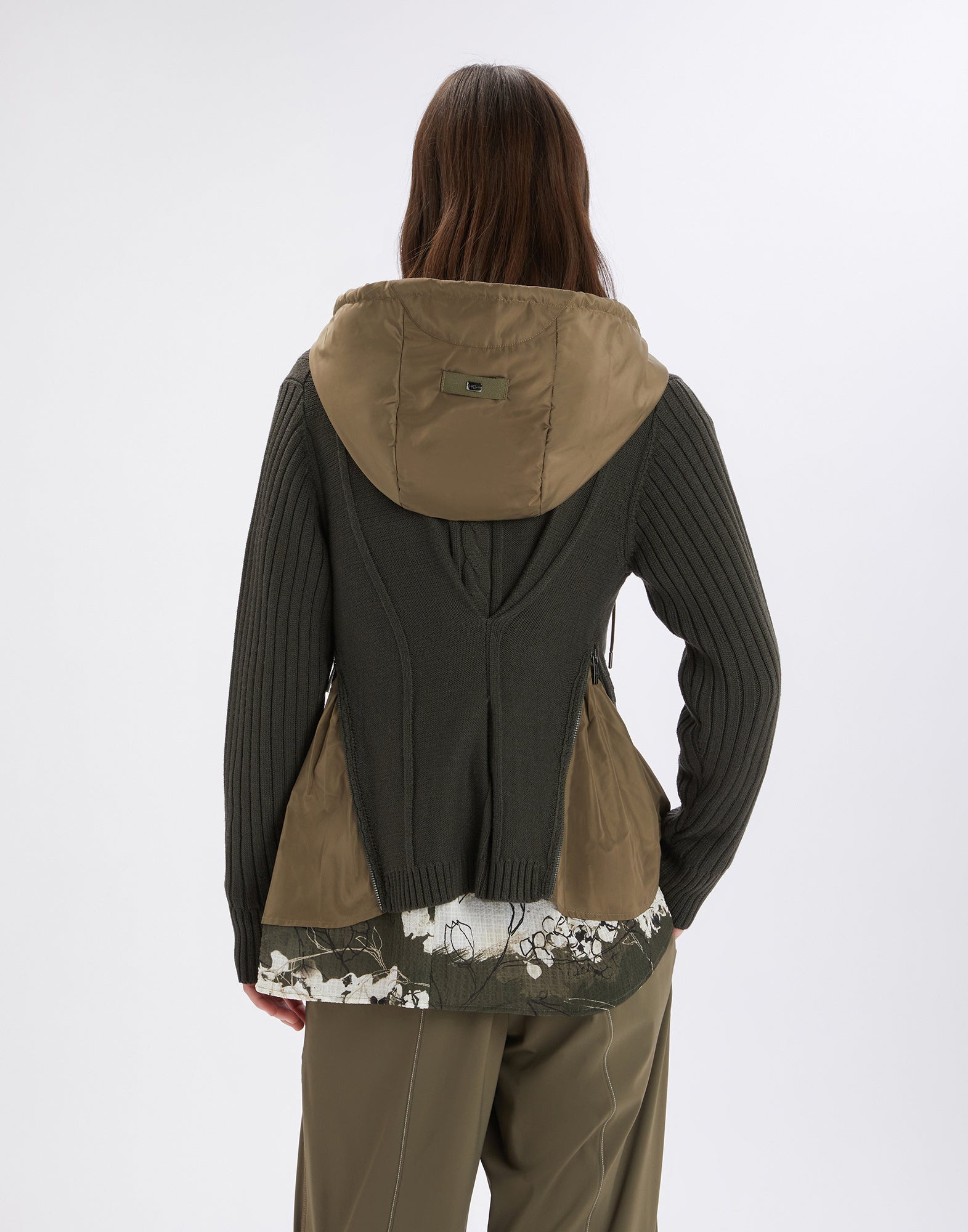 Back view of a woman wearing the FREIZE cardigan, showing nylon inserts at the sides, a detachable hood, and asymmetric pleat.