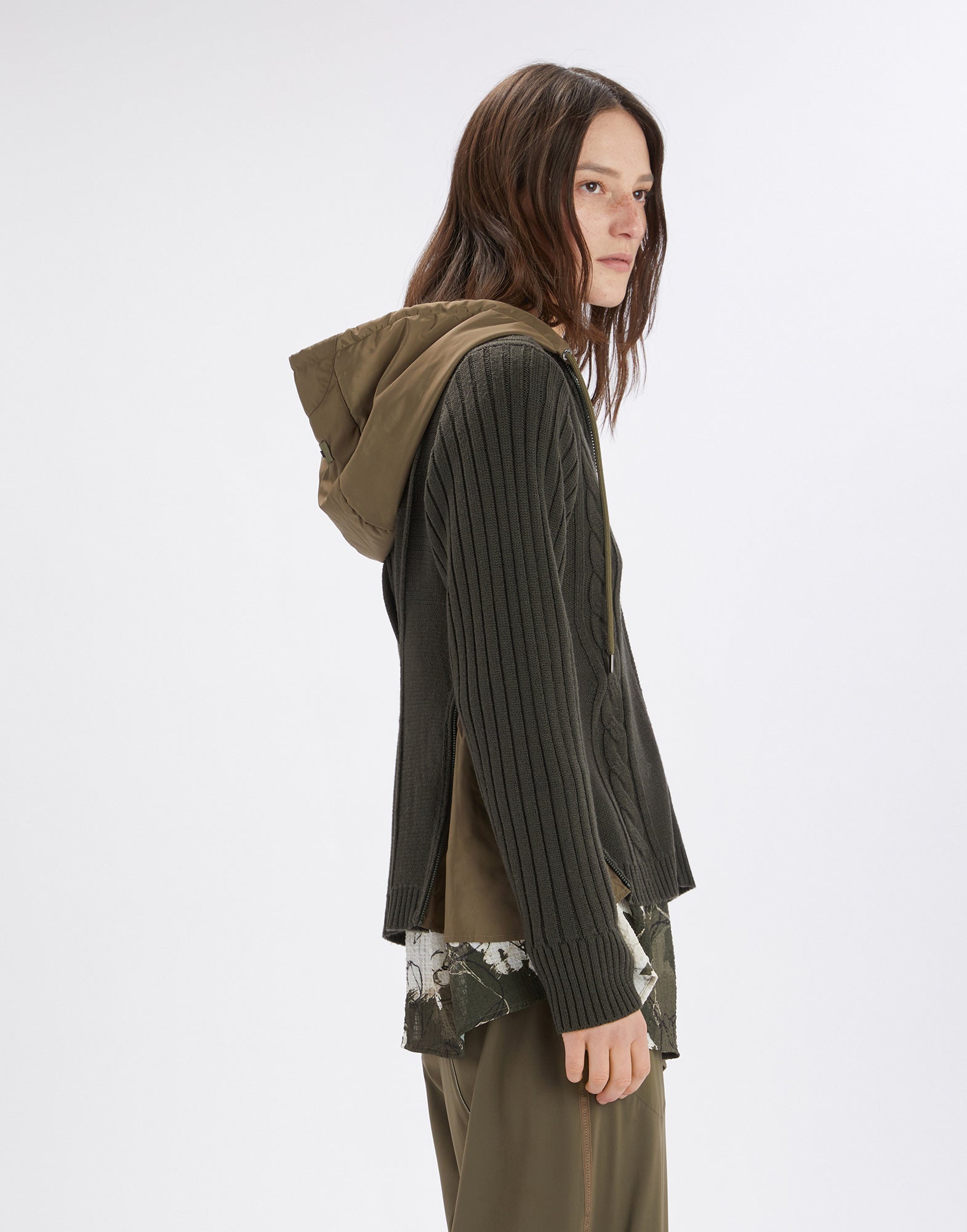 Side view of a person wearing the FREIZE cardigan, showing a cable and rib knit pattern with olive detachable hood and side nylon inserts.