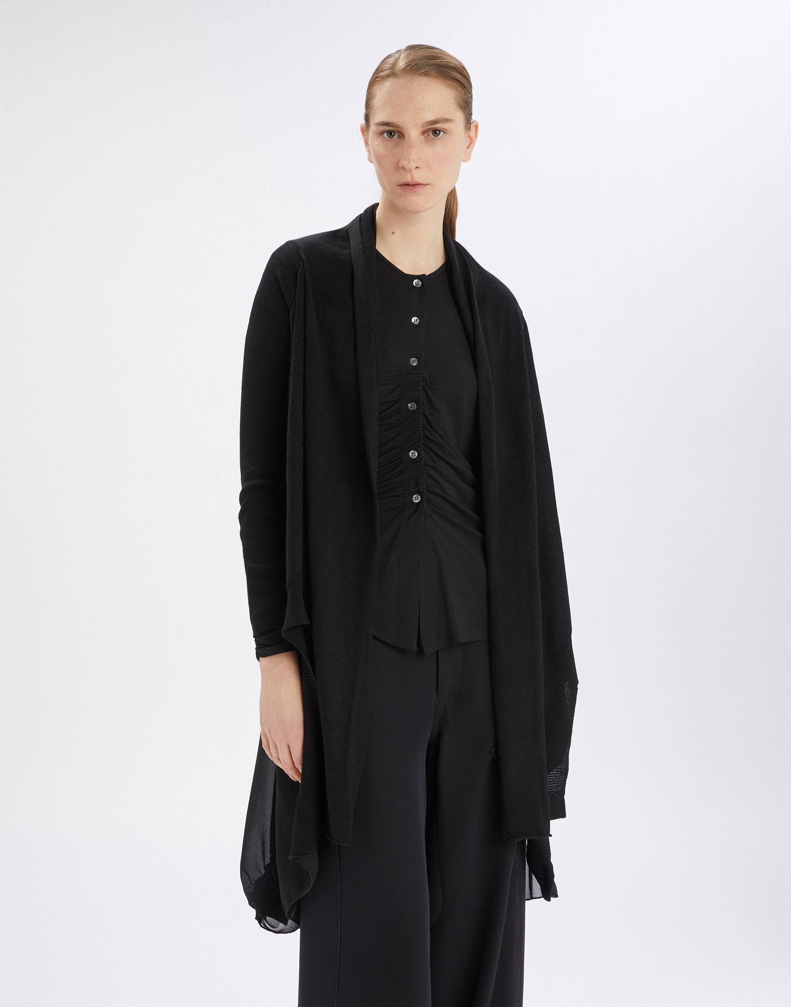 Frontalansicht: Eine Frau trägt den Cardigan SLIVER in Schwarz, locker geschnitten ohne Verschluss, mit langen Rippenärmeln und unregelmäßigem Saum über schwarzer Kleidung.