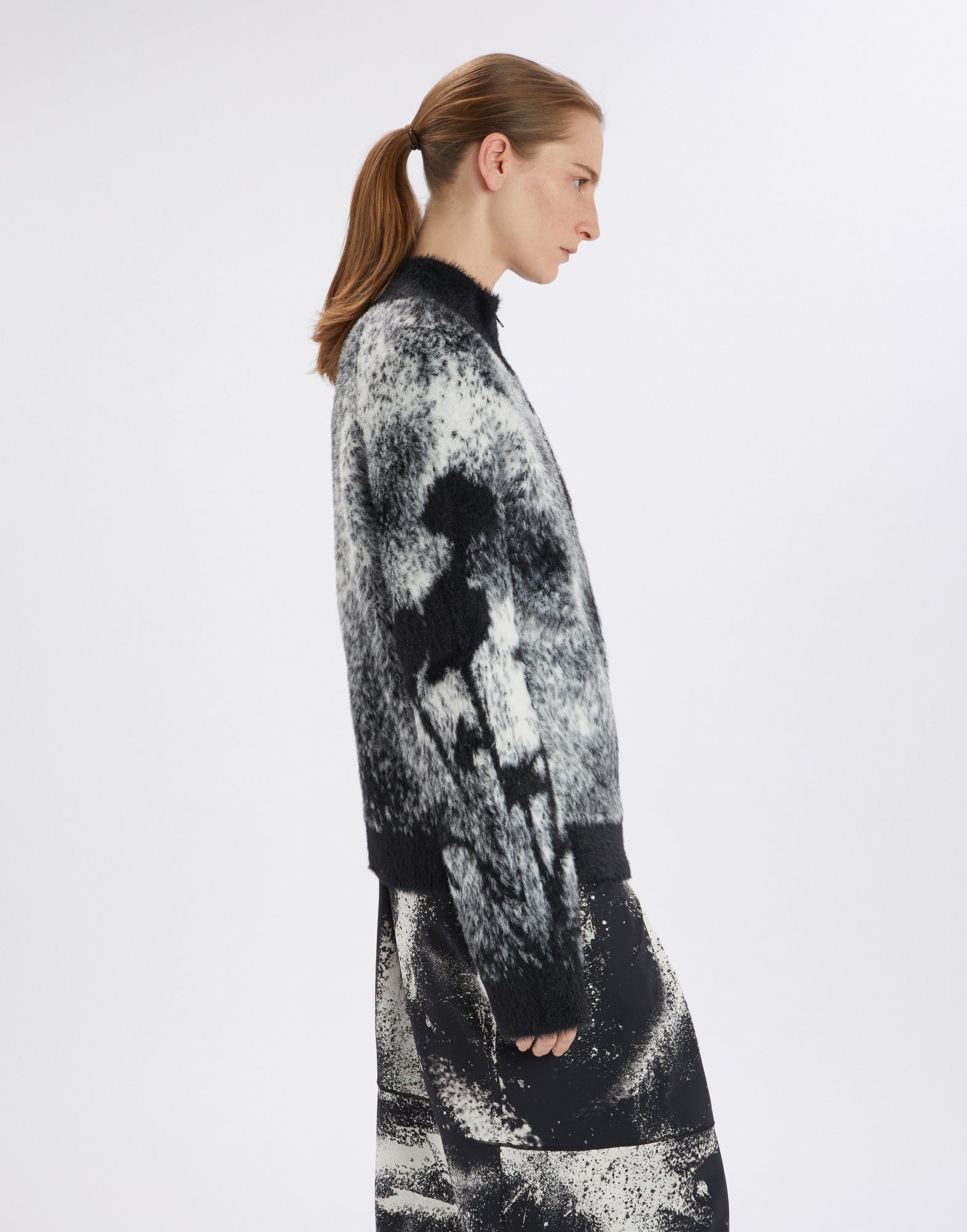 Seitenansicht eines Models, das den SUMMON UP Cardigan trägt. Der Cardigan hat ein bicolor Jacquard-Muster, florales Detail am rechten Ärmel und unifarbene Abschlüsse.